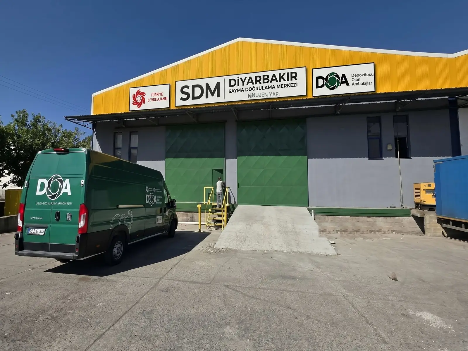Diyarbakır Sayma Doğrulama Merkezi cephesi; Türkiye Çevre Ajansı, SDM ve DOA tabelaları, önünde DOA logolu toplama aracı ve sevkiyat rampası